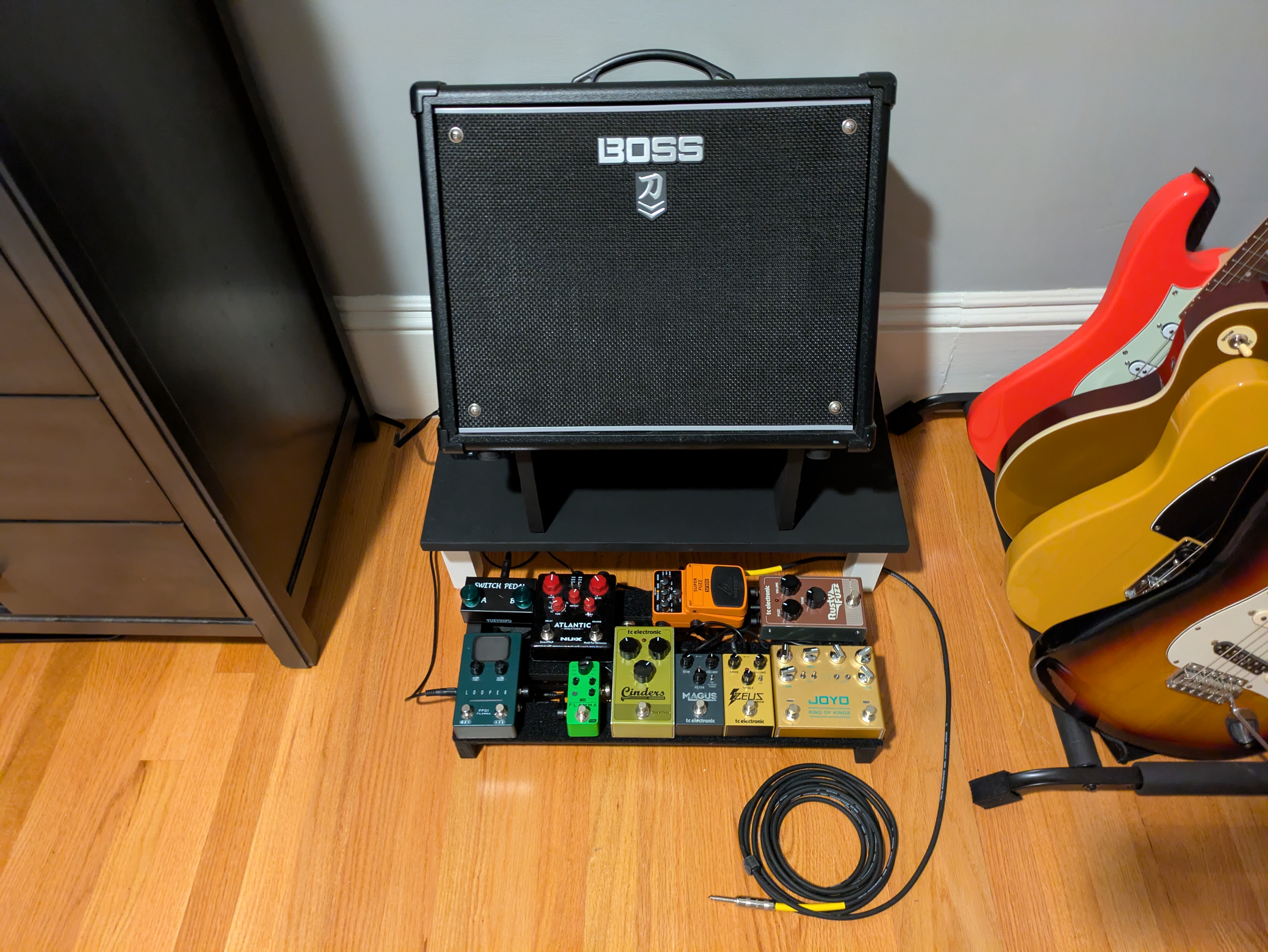 The pedalboard in front of the platform that has the amp in the tilt-back stand on top of it
